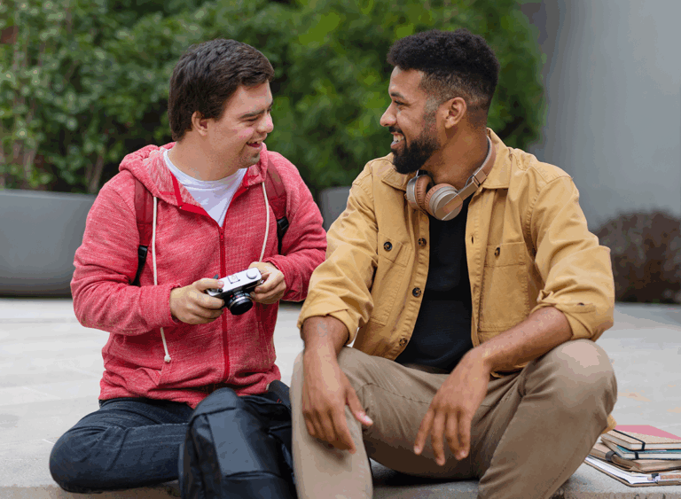A happy young man with Down syndrome and mentoring friend sitting outdoors